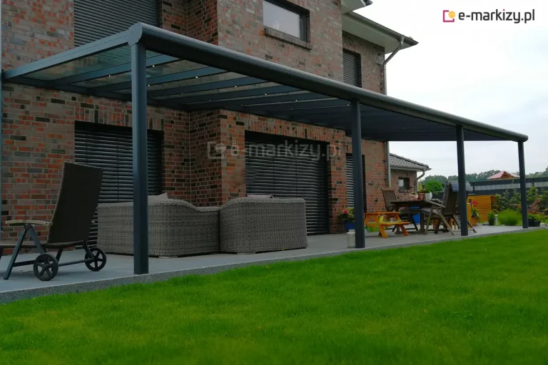 Zdjęcie prezentuje wielkogabarytowe, 12-metrowe zadaszenie tarasu model Terano, wykonane z profili aluminiowych w kolorze antracytowym. Konstrukcja jest zamontowana bezpośrednio do elewacji piętrowego budynku z wielokolorowej cegły klinkierowej. Dach zadaszenia jest przeszklony, a pod krokwiami zamontowano punktowe oświetlenie LED, które doświetla przestrzeń tarasu. Całość opiera się na czterech solidnych słupach nośnych o przekroju kwadratowym, rozmieszczonych wzdłuż krawędzi betonowej wylewki tarasowej. Przednia belka konstrukcyjna posiada zaokrąglony profil i pełni funkcję rynny odprowadzającej wodę. Na tarasie pod zadaszeniem znajdują się meble ogrodowe, w tym duże fotele technorattanowe w odcieniu szarości, drewniany zestaw mebli jadalnianych oraz leżak. Okna budynku wyposażone są w czarne rolety zewnętrzne typu żaluzje fasadowe. Przed tarasem rozciąga się zadbany, zielony trawnik, a w tle widoczne są fragmenty sąsiedniej zabudowy pod pochmurnym niebem.