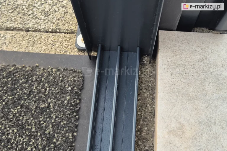 Trzytorowa prowadnica dolna do systemu szklanych ścian przesuwnych Miros z widocznym w górnej części profilem doszczelniającym U, przymocowanym do profilu zadaszenia tarasu. Po lewej stronie prowadnicy widoczna wycieraczka w odcieniach grafitu, po prawej płyta tarasowa w odcieniach beżu i szarości.