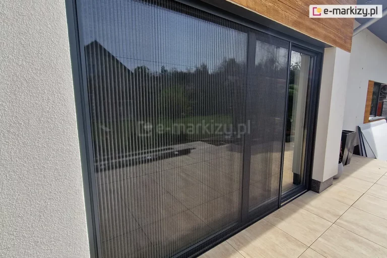 Moskitiera Top Zag o szerokości 4 m zamocowana na dużym oknie tarasowym z częściowo odsuniętą siatką. Na tarasie położone duże płytki imitujące drewno. W tle donice, płyty ustawione pod ścianą oraz okno pokojowe.