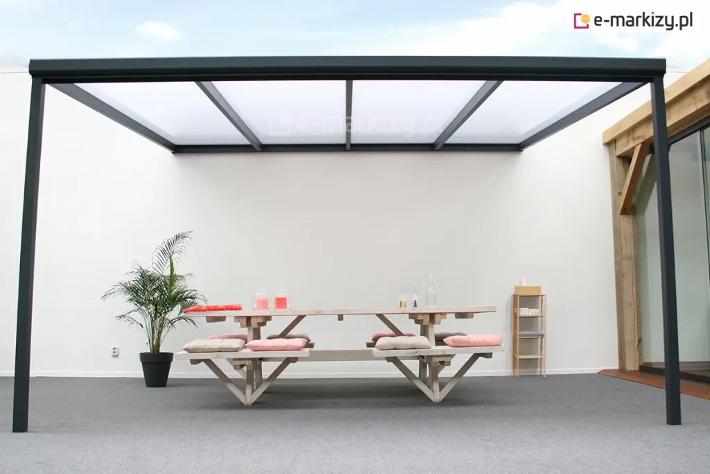 Zdjęcie prezentuje nowoczesne, 4-metrowe zadaszenie tarasowe Nebbiolo Deponti o minimalistycznej konstrukcji aluminiowej w kolorze ciemnoantracytowym. Konstrukcja składa się z dwóch smukłych słupów nośnych oraz ramy dachowej z równolegle rozmieszczonymi krokiewkami, w których osadzono płyty z poliwęglanu komorowego o mlecznym wybarwieniu. Zadaszenie jest zamontowane w przestrzeni o białych, gładkich ścianach, co podkreśla jego geometryczną formę. Pod wiatą znajduje się duży stół piknikowy zintegrowany z ławami, wykonany z jasnego drewna i wyposażony w miękkie poduszki w kolorze bladoróżowym. Aranżację uzupełnia wysoka roślina doniczkowa w czarnym pojemniku oraz drewniany regał stojący przy ścianie. Po prawej stronie widoczny jest fragment przeszklenia oraz drewnianej konstrukcji budynku, a podłoże stanowi jednolita, szara nawierzchnia.