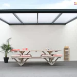 Zdjęcie prezentuje nowoczesne, 4-metrowe zadaszenie tarasowe Nebbiolo Deponti o minimalistycznej konstrukcji aluminiowej w kolorze ciemnoantracytowym. Konstrukcja składa się z dwóch smukłych słupów nośnych oraz ramy dachowej z równolegle rozmieszczonymi krokiewkami, w których osadzono płyty z poliwęglanu komorowego o mlecznym wybarwieniu. Zadaszenie jest zamontowane w przestrzeni o białych, gładkich ścianach, co podkreśla jego geometryczną formę. Pod wiatą znajduje się duży stół piknikowy zintegrowany z ławami, wykonany z jasnego drewna i wyposażony w miękkie poduszki w kolorze bladoróżowym. Aranżację uzupełnia wysoka roślina doniczkowa w czarnym pojemniku oraz drewniany regał stojący przy ścianie. Po prawej stronie widoczny jest fragment przeszklenia oraz drewnianej konstrukcji budynku, a podłoże stanowi jednolita, szara nawierzchnia.