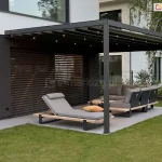 Przydomowy taras z eleganckim zadaszeniem pergolą Square Flat. Konstrukcja wykonana jest z ciemnoszarego metalu, z prostymi słupami i dachem z poziomych belek, pomiędzy którymi rozpięta jest jasnoszara tkanina. W belkach osadzone są subtelne punkty świetlne LED, dające ciepłe światło, które oświetla przestrzeń wypoczynkową pod pergolą. Na pierwszym planie znajduje się nowoczesny leżak z tapicerowanym siedziskiem w odcieniu jasnej szarości, umieszczony na drewnianej ramie z czarnymi nogami. Na leżaku leży ozdobna, żółta poduszka. W głębi widać narożną sofę ogrodową utrzymaną w tym samym stylu i kolorystyce, z dużymi poduszkami i drewnianą podstawą. Między meblami rozmieszczone są niskie stoliki kawowe. Cała przestrzeń osadzona jest na szarej, płytkowanej powierzchni tarasowej, otoczonej intensywnie zielonym trawnikiem. W tle widoczne są poziome żaluzje zewnętrzne, zieleń ogrodu oraz fragment jasnej elewacji budynku i sąsiedniej zabudowy. Kompozycja oddaje poczucie luksusu, nowoczesności i komfortu.