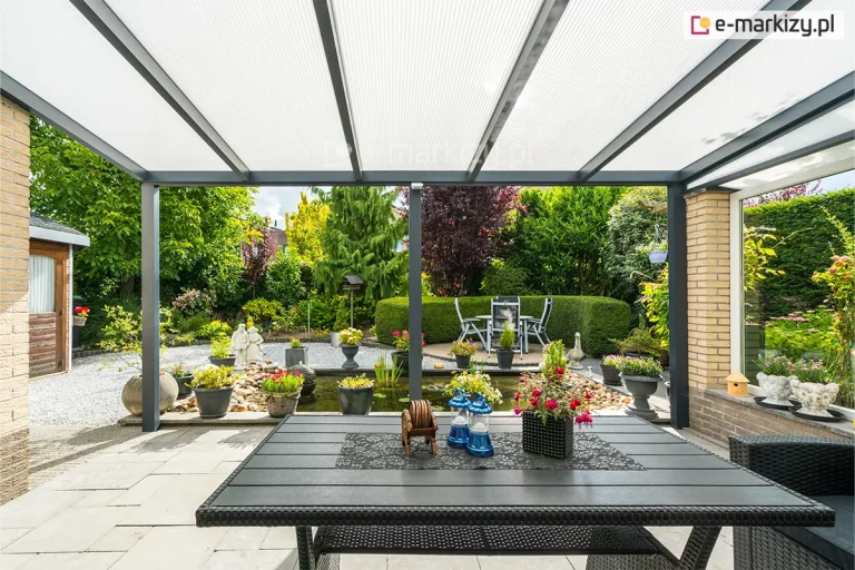 Szeroki widok spod zadaszenia tarasowego na pięknie zagospodarowany ogród z oczkiem wodnym. Konstrukcja dachu opiera się na antracytowych profilach aluminiowych, wypełnionych panelami z poliwęglanu komorowego w kolorze mlecznym, który zapewnia jasne i rozproszone światło. Na tarasie, wyłożonym jasnymi płytami, ustawiono duży stół ogrodowy z technorattanu, ozdobiony kwiatami i dekoracyjnymi lampionami. W tle widać bogatą roślinność: przystrzyżone żywopłoty, krzewy ozdobne, kwiaty w donicach oraz kamieniste ścieżki otaczające staw z fontanną.