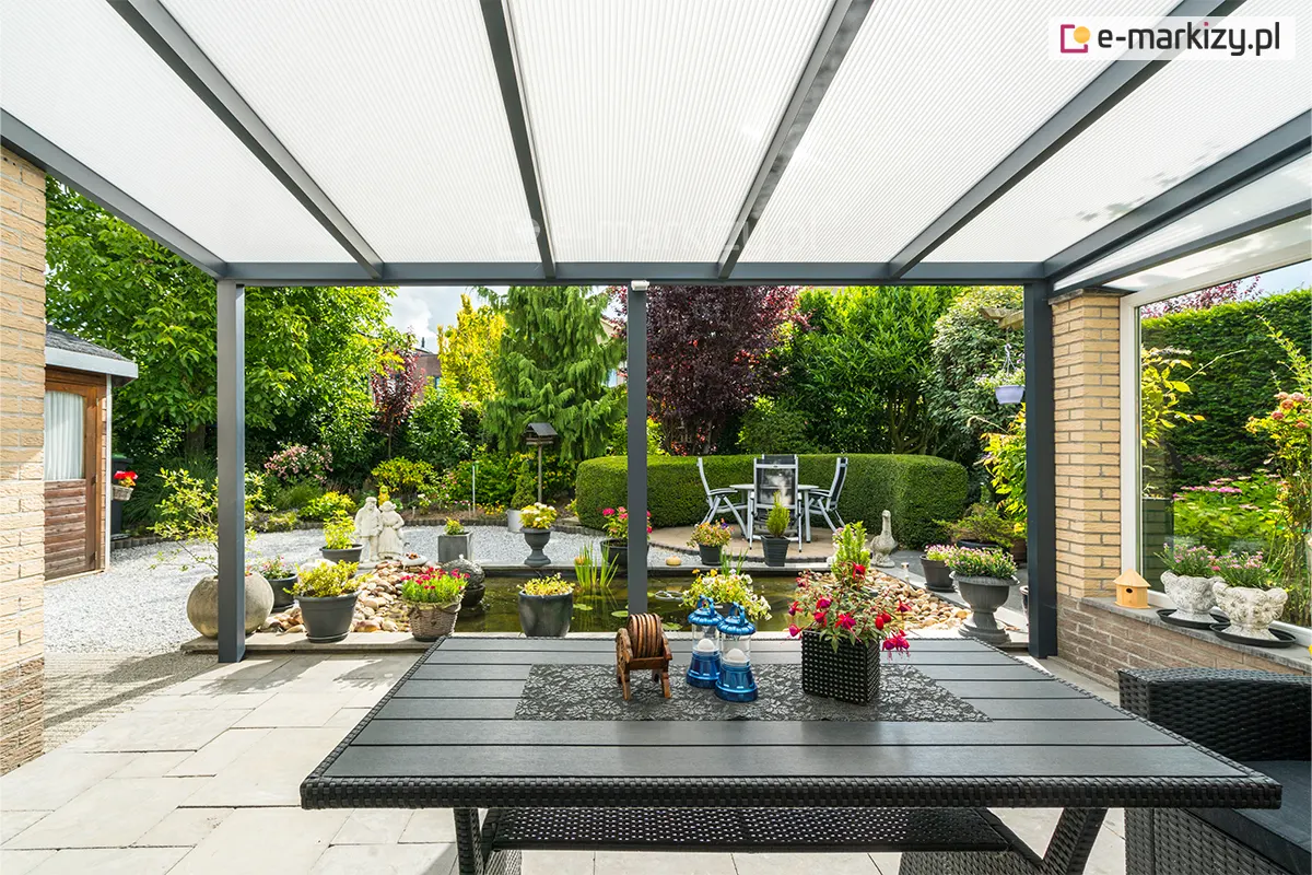 Szeroki widok spod zadaszenia tarasowego na pięknie zagospodarowany ogród z oczkiem wodnym. Konstrukcja dachu opiera się na antracytowych profilach aluminiowych, wypełnionych panelami z poliwęglanu komorowego w kolorze mlecznym, który zapewnia jasne i rozproszone światło. Na tarasie, wyłożonym jasnymi płytami, ustawiono duży stół ogrodowy z technorattanu, ozdobiony kwiatami i dekoracyjnymi lampionami. W tle widać bogatą roślinność: przystrzyżone żywopłoty, krzewy ozdobne, kwiaty w donicach oraz kamieniste ścieżki otaczające staw z fontanną.