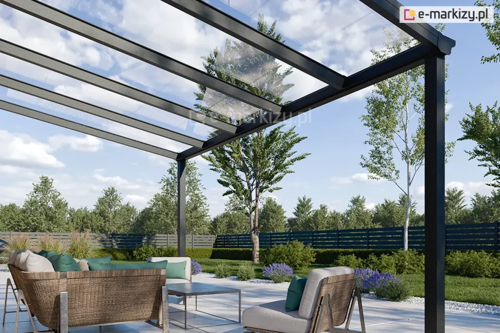 Zdjęcie prezentuje zbliżenie na konstrukcję nowoczesnego, 5-metrowego zadaszenia tarasowego Terano, wykonanego z profili aluminiowych w kolorze antracytowym. Widok z perspektywy żabiej podkreśla transparentność dachu, który został wypełniony panelami ze szkła bezpiecznego, opartymi na systemowych krokwiach aluminiowych. Konstrukcja wspiera się na pionowych słupach o kwadratowym przekroju, a przednia belka wieńcząca zintegrowana jest z systemem odprowadzania wody deszczowej. Pod zadaszeniem zaaranżowano nowoczesną strefę wypoczynkową z meblami ogrodowymi o konstrukcji drewniano-metalowej, wyposażonymi w jasnoszare siedziska i kontrastujące, zielone poduszki. Nawierzchnia tarasu wykonana jest z jasnych płyt wielkoformatowych, otoczonych opaską z białego grysiku. W tle widoczny jest estetyczny ogród z zielonym trawnikiem, ozdobnymi krzewami, lawendą oraz ażurowym, ciemnym ogrodzeniem frontowym, za którym rosną wysokie drzewa liściaste pod błękitnym niebem z rozproszonymi chmurami.
