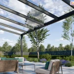 Zdjęcie prezentuje zbliżenie na konstrukcję nowoczesnego, 5-metrowego zadaszenia tarasowego Terano, wykonanego z profili aluminiowych w kolorze antracytowym. Widok z perspektywy żabiej podkreśla transparentność dachu, który został wypełniony panelami ze szkła bezpiecznego, opartymi na systemowych krokwiach aluminiowych. Konstrukcja wspiera się na pionowych słupach o kwadratowym przekroju, a przednia belka wieńcząca zintegrowana jest z systemem odprowadzania wody deszczowej. Pod zadaszeniem zaaranżowano nowoczesną strefę wypoczynkową z meblami ogrodowymi o konstrukcji drewniano-metalowej, wyposażonymi w jasnoszare siedziska i kontrastujące, zielone poduszki. Nawierzchnia tarasu wykonana jest z jasnych płyt wielkoformatowych, otoczonych opaską z białego grysiku. W tle widoczny jest estetyczny ogród z zielonym trawnikiem, ozdobnymi krzewami, lawendą oraz ażurowym, ciemnym ogrodzeniem frontowym, za którym rosną wysokie drzewa liściaste pod błękitnym niebem z rozproszonymi chmurami.