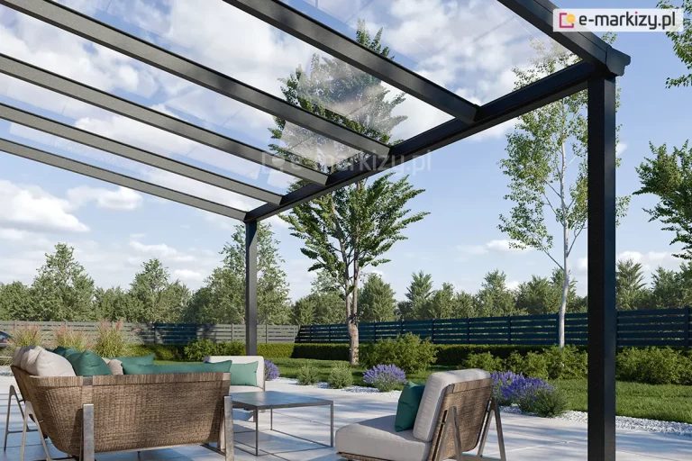 Zdjęcie prezentuje zbliżenie na konstrukcję nowoczesnego, 5-metrowego zadaszenia tarasowego Terano, wykonanego z profili aluminiowych w kolorze antracytowym. Widok z perspektywy żabiej podkreśla transparentność dachu, który został wypełniony panelami ze szkła bezpiecznego, opartymi na systemowych krokwiach aluminiowych. Konstrukcja wspiera się na pionowych słupach o kwadratowym przekroju, a przednia belka wieńcząca zintegrowana jest z systemem odprowadzania wody deszczowej. Pod zadaszeniem zaaranżowano nowoczesną strefę wypoczynkową z meblami ogrodowymi o konstrukcji drewniano-metalowej, wyposażonymi w jasnoszare siedziska i kontrastujące, zielone poduszki. Nawierzchnia tarasu wykonana jest z jasnych płyt wielkoformatowych, otoczonych opaską z białego grysiku. W tle widoczny jest estetyczny ogród z zielonym trawnikiem, ozdobnymi krzewami, lawendą oraz ażurowym, ciemnym ogrodzeniem frontowym, za którym rosną wysokie drzewa liściaste pod błękitnym niebem z rozproszonymi chmurami.