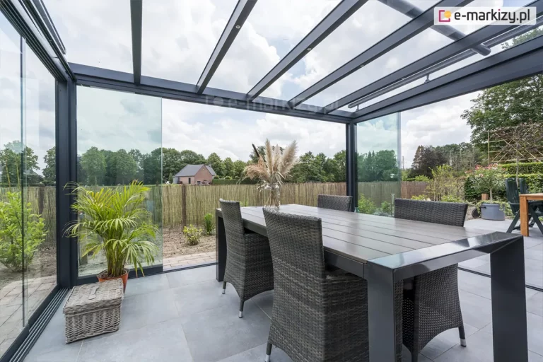 Antracytowe zadaszenie tarasowe marki Deponti o konstrukcji aluminiowej, wyposażone w szklany dach oraz system przesuwnych szyb bocznych, tworzące zamknięty ogród zimowy. Konstrukcja dachu składa się z ciemnych profili utrzymujących transparentne tafle szkła, przez które widoczne jest zachmurzone niebo. Pod zadaszeniem znajduje się strefa jadalniana z dużym stołem o szarym blacie oraz czterema krzesłami wykonanymi z ciemnego technorattanu. Wnętrze jest minimalistyczne, z jasnoszarą podłogą z płyt tarasowych, na której stoi pleciony kosz oraz donica z zieloną palmą. Szklane panele boczne są częściowo rozsunięte, otwierając widok na ogród otoczony słomianym płotem, niską roślinnością oraz drzewami w oddali. W tle, poza terenem posesji, widoczny jest dom jednorodzinny z ceglaną elewacją.