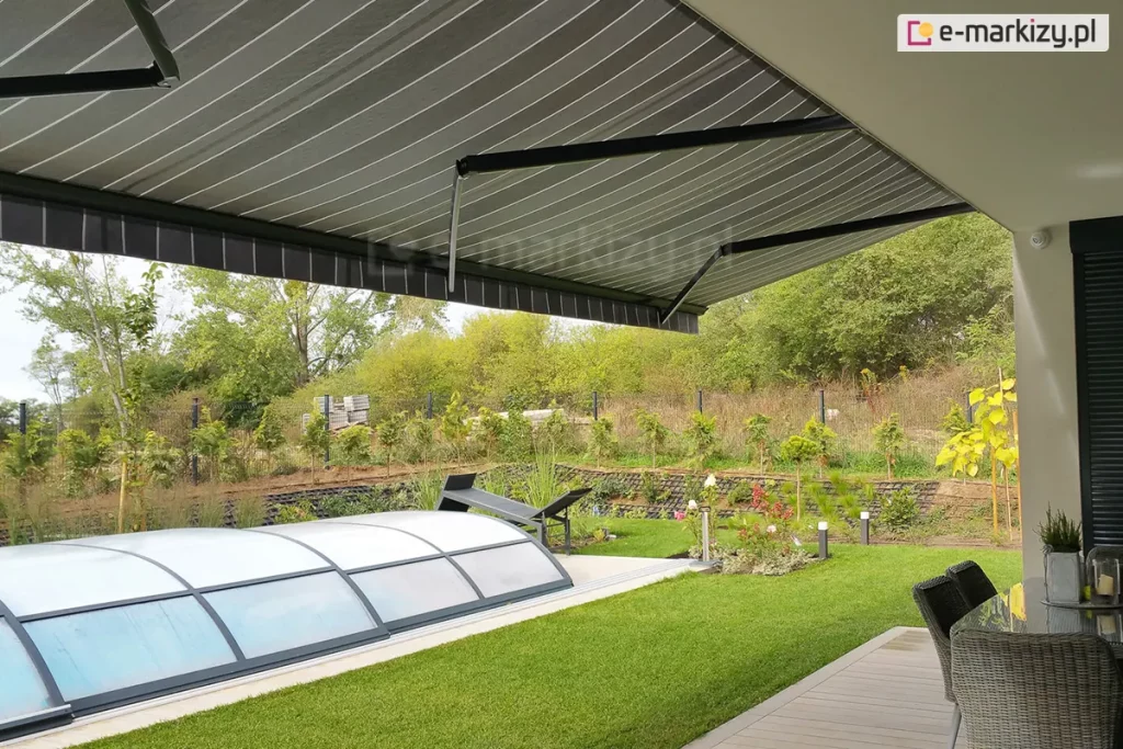 siedmiometrowa markiza Adagio MOL z trzema ramionami w kolorze antracytowym z rozwiniętym nad tarasem i trawnikiem poszyciem w szare i białe pasy. Pod markizą widoczny częściowo drewniany taras ze stołem przykrytym szkłem i krzesłami z rattanu. W ogrodzie kopuła krytego basenu oraz zadbany trawnik, a w tle liczne kwiaty i rośliny. Ogród otaczają drzewa i krzewy o zielonych liściach.