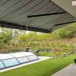 siedmiometrowa markiza Adagio MOL z trzema ramionami w kolorze antracytowym z rozwiniętym nad tarasem i trawnikiem poszyciem w szare i białe pasy. Pod markizą widoczny częściowo drewniany taras ze stołem przykrytym szkłem i krzesłami z rattanu. W ogrodzie kopuła krytego basenu oraz zadbany trawnik, a w tle liczne kwiaty i rośliny. Ogród otaczają drzewa i krzewy o zielonych liściach.