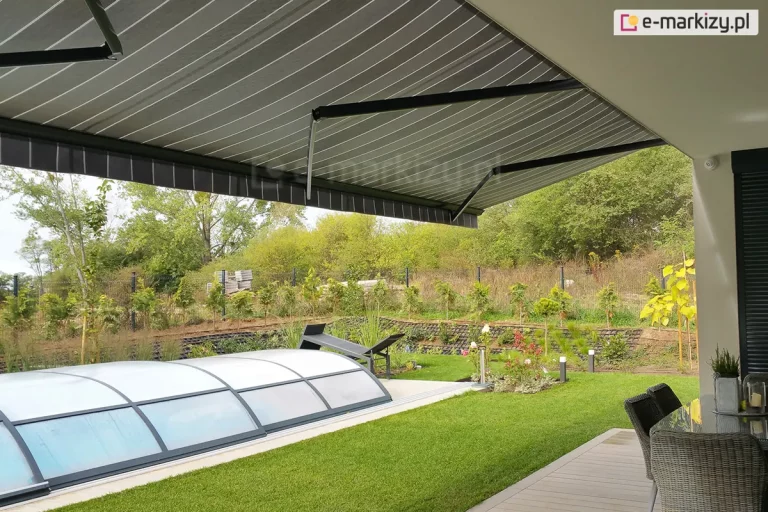 siedmiometrowa markiza Adagio MOL z trzema ramionami w kolorze antracytowym z rozwiniętym nad tarasem i trawnikiem poszyciem w szare i białe pasy. Pod markizą widoczny częściowo drewniany taras ze stołem przykrytym szkłem i krzesłami z rattanu. W ogrodzie kopuła krytego basenu oraz zadbany trawnik, a w tle liczne kwiaty i rośliny. Ogród otaczają drzewa i krzewy o zielonych liściach.