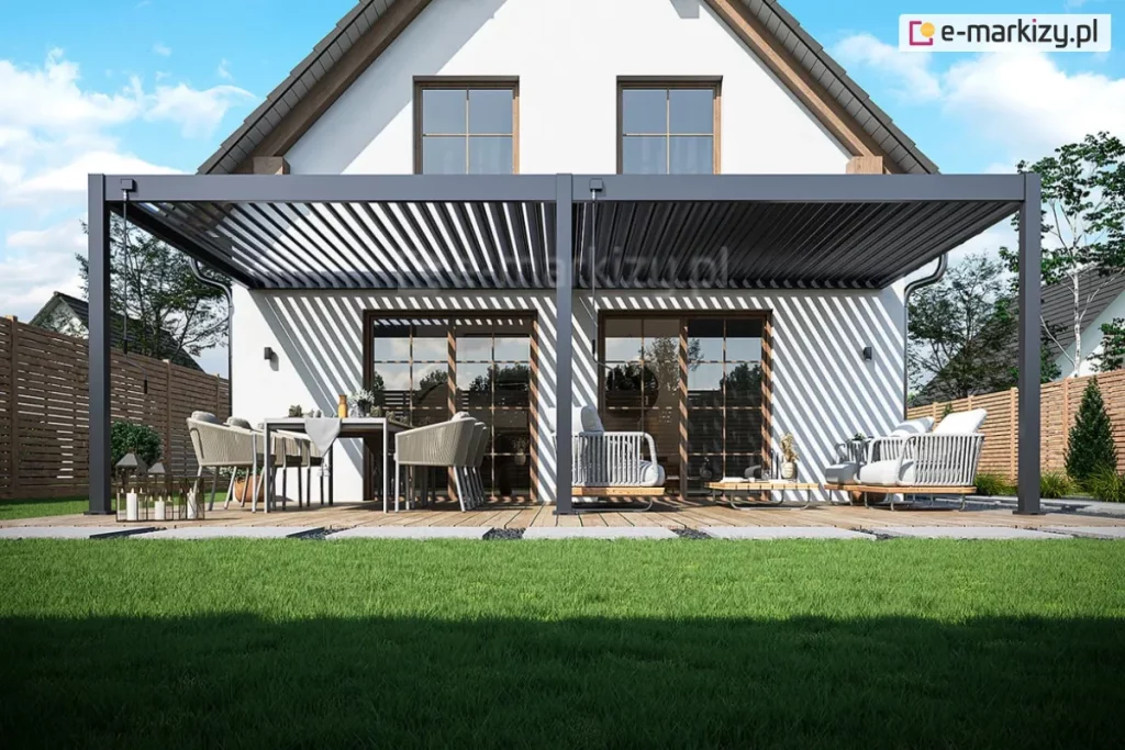 Przyścienna pergola lamelowa Rubio Soft o konstrukcji aluminiowej w kolorze antracytowym zamontowana do ściany na białej elewacji piętrowego domu jednorodzinnego. Zadaszenie pergoli składa się z ruchomych lameli ustawionych pod kątem, które rzucają charakterystyczny liniowy cień na ścianę budynku oraz deskowanie tarasu. Konstrukcja opiera się na trzech masywnych pionowych słupach i przedniej belce poprzecznej. Pod pergolą znajduje się luksusowo urządzona strefa wypoczynkowa na drewnianym tarasie. Po lewej stronie ustawiono prostokątny stół z szarym obrusem oraz sześć nowoczesnych foteli z technorattanu z jasnymi poduszkami. Na stole znajdują się elementy zastawy oraz dekoracje. Po prawej stronie umieszczono zestaw mebli ogrodowych składający się z sofy i foteli o identycznym designie z grubymi szarymi poduchami oraz niskiego stolika kawowego. Taras otoczony jest zadbanym trawnikiem, a w tle widać drewniane ogrodzenie lamelowe oddzielające posesję od sąsiednich działek. Dom posiada dwuspadowy dach o ciemnym pokryciu oraz duże przeszklenia w postaci drzwi tarasowych ze szprosami i dwóch symetrycznych okien na poddaszu. Całość przedstawiona jest w pełnym słońcu przy bezchmurnym niebie, co podkreśla nowoczesną architekturę budynku i funkcjonalność systemu osłonowego.