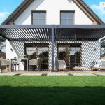Przyścienna pergola lamelowa Rubio Soft o konstrukcji aluminiowej w kolorze antracytowym zamontowana do ściany na białej elewacji piętrowego domu jednorodzinnego. Zadaszenie pergoli składa się z ruchomych lameli ustawionych pod kątem, które rzucają charakterystyczny liniowy cień na ścianę budynku oraz deskowanie tarasu. Konstrukcja opiera się na trzech masywnych pionowych słupach i przedniej belce poprzecznej. Pod pergolą znajduje się luksusowo urządzona strefa wypoczynkowa na drewnianym tarasie. Po lewej stronie ustawiono prostokątny stół z szarym obrusem oraz sześć nowoczesnych foteli z technorattanu z jasnymi poduszkami. Na stole znajdują się elementy zastawy oraz dekoracje. Po prawej stronie umieszczono zestaw mebli ogrodowych składający się z sofy i foteli o identycznym designie z grubymi szarymi poduchami oraz niskiego stolika kawowego. Taras otoczony jest zadbanym trawnikiem, a w tle widać drewniane ogrodzenie lamelowe oddzielające posesję od sąsiednich działek. Dom posiada dwuspadowy dach o ciemnym pokryciu oraz duże przeszklenia w postaci drzwi tarasowych ze szprosami i dwóch symetrycznych okien na poddaszu. Całość przedstawiona jest w pełnym słońcu przy bezchmurnym niebie, co podkreśla nowoczesną architekturę budynku i funkcjonalność systemu osłonowego.