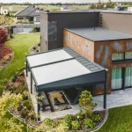 Widok z lotu ptaka na nowoczesną, dwumodułową pergolę tarasową Elegancy Tarasola o konstrukcji aluminiowej w kolorze antracytowym, zamontowaną do elewacji budynku o surowej, industrialnej stylistyce. Zadaszenie składa się z dwóch niezależnych sekcji z jasną, napiętą tkaniną osłonową, która skutecznie kontrastuje z ciemną ramą systemu. Pod pergolą, na tarasie wyłożonym wielkoformatowymi, jasnoszarymi płytami, znajduje się strefa relaksu wyposażona w ciemny narożnik ogrodowy, stolik z drewnianym blatem oraz profesjonalny grill gazowy. Elewacja budynku łączy ciemny tynk strukturalny z panelami o efekcie rdzy (corten), co podkreśla nowoczesny charakter inwestycji. Całość otacza starannie zaaranżowany ogród z zielonym trawnikiem, półkolistymi rabatami wysypanymi dekoracyjną korą oraz różnorodnymi nasadzeniami krzewów i drzew iglastych, a w oddali widoczna jest sąsiednia zabudowa jednorodzinna pod zachmurzonym niebem.