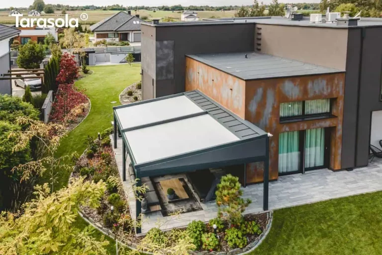 Widok z lotu ptaka na nowoczesną, dwumodułową pergolę tarasową Elegancy Tarasola o konstrukcji aluminiowej w kolorze antracytowym, zamontowaną do elewacji budynku o surowej, industrialnej stylistyce. Zadaszenie składa się z dwóch niezależnych sekcji z jasną, napiętą tkaniną osłonową, która skutecznie kontrastuje z ciemną ramą systemu. Pod pergolą, na tarasie wyłożonym wielkoformatowymi, jasnoszarymi płytami, znajduje się strefa relaksu wyposażona w ciemny narożnik ogrodowy, stolik z drewnianym blatem oraz profesjonalny grill gazowy. Elewacja budynku łączy ciemny tynk strukturalny z panelami o efekcie rdzy (corten), co podkreśla nowoczesny charakter inwestycji. Całość otacza starannie zaaranżowany ogród z zielonym trawnikiem, półkolistymi rabatami wysypanymi dekoracyjną korą oraz różnorodnymi nasadzeniami krzewów i drzew iglastych, a w oddali widoczna jest sąsiednia zabudowa jednorodzinna pod zachmurzonym niebem.