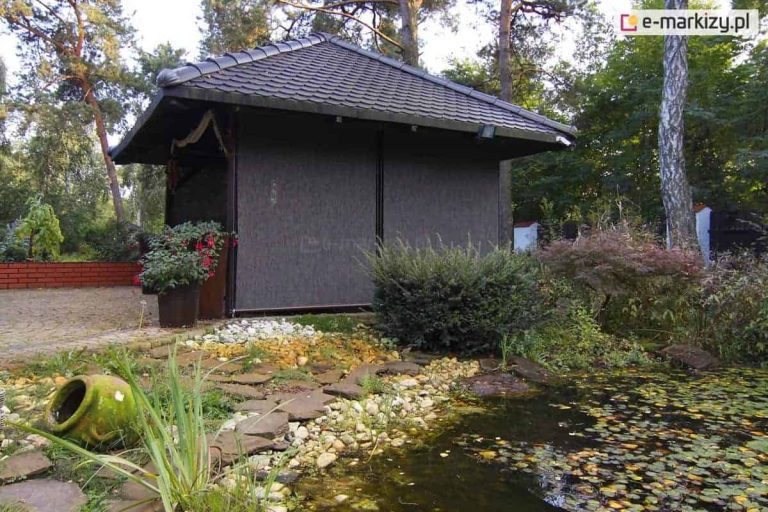 Altana ogrodowa z dachem pokrytym dachówką, wyposażona w dwie zewnętrzne rolety screenowe w ciemnym kolorze, opuszczone do samego dołu. Rolety zamontowane są na bocznej ścianie altany, tworząc osłonę przeciwsłoneczną i prywatnościową. Budynek otoczony bujną roślinnością, z przodu widoczny skalniak oraz oczko wodne z kamieniami, roślinami wodnymi i ceramicznym naczyniem. Tło stanowi gęsty las iglasto-liściasty.