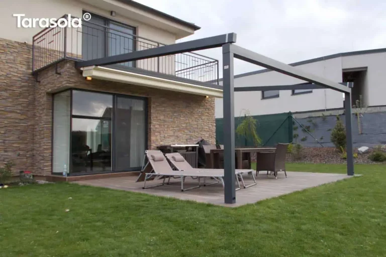 Nowoczesna aluminiowa pergola tarasowa Elegancy Tarasola o konstrukcji przyściennej w kolorze antracytowym, z całkowicie zwiniętym zadaszeniem tkaninowym ukrytym pod systemowym daszkiem osłonowym. Stelaż składa się z prowadnic dachowych zamontowanych do elewacji oraz dwóch przednich słupów wsporczych o kwadratowym przekroju, posadowionych na tarasie wyłożonym płytkami imitującymi drewno. Elewacja budynku na parterze wykończona jest ozdobnym kamieniem w odcieniach beżu i brązu, z dużym przeszkleniem prowadzącym do wnętrza domu. Na tarasie znajdują się dwa beżowe leżaki ogrodowe z zagłówkami oraz stół jadalniany z krzesłami w tle. Powyżej widoczny jest balkon z metalową balustradą. Otoczenie stanowi zadbany trawnik, niskie nasadzenia na rabacie z kory oraz zielone ogrodzenie siatkowe, za którym znajduje się sąsiedni budynek mieszkalny o jasnej elewacji.