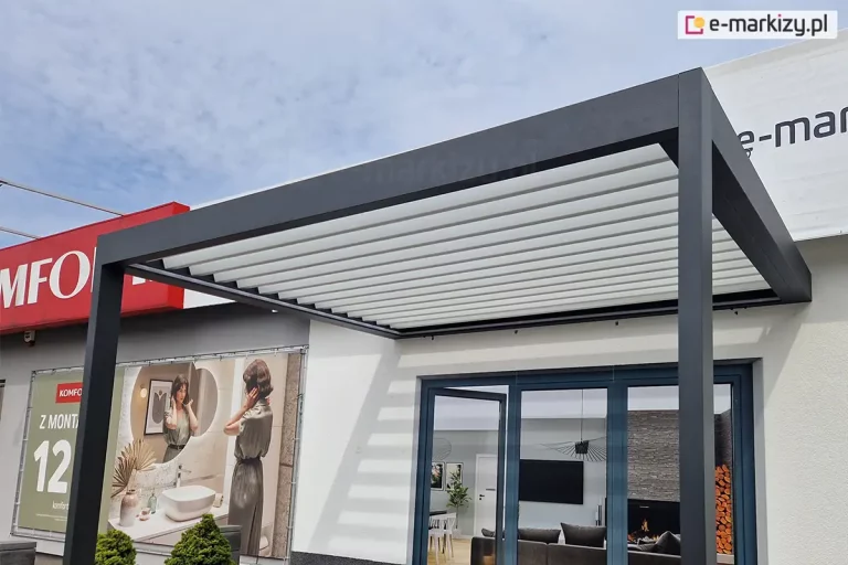 Nowoczesna pergola tarasowa przyścienna SB400 Selt o konstrukcji aluminiowej w kolorze ciemnego antracytu, zamontowana do elewacji budynku. Zadaszenie bioklimatyczne składa się z białych, ruchomych lameli, które są uchylone, rzucają charakterystyczny paskowany cień na ścianę. Konstrukcja wsparta jest na dwóch masywnych, kwadratowych profilach przednich. Pergola osłania wejście do budynku z dużymi, przeszklonymi drzwiami tarasowymi, w których odbija się wnętrze salonu z kanapą i kominkiem. Na elewacji obok pergoli widoczny jest duży baner reklamowy salonu łazienek oraz fragment czerwonego szyldu. W lewym dolnym rogu zdjęcia znajdują się dwa małe, przycięte krzewy iglaste. Niebo w tle jest lekko zachmurzone, w kolorze jasnoszarym.