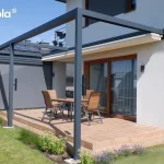 Zdjęcie przedstawia aluminiową pergolę tarasową Tarasola Flat o konstrukcji przyściennej w kolorze ciemnego antracytu, zamontowaną przy nowoczesnym domu jednorodzinnym. Zadaszenie tkaninowe w kolorze kremowym jest w tym momencie całkowicie zwinięte pod systemowy daszek ochronny, co odsłania pełną ramę konstrukcji oraz przestrzeń tarasu. Na jednym z boków pergoli zainstalowany jest pionowy refleksol Zip z tkaniną typu screen w kolorze szarym, która jest w pełni opuszczona, zapewniając osłonę boczną. Taras został wyłożony płytkami w ciepłym odcieniu brązu i wyposażony w drewniany stół oraz cztery krzesła z tekstylnym obiciem. Na słupach konstrukcyjnych widoczne są zamontowane akcesoria, prawdopodobnie kinkiety lub głośniki. Otoczenie stanowi zadbany trawnik z niskimi nasadzeniami lawendy i traw ozdobnych wzdłuż krawędzi podestu. Elewacja budynku jest jasna, z dużymi przesuwnymi drzwiami tarasowymi w drewnianej ramie, a w tle widoczne są sąsiednie zabudowania z panelami fotowoltaicznymi na dachu.