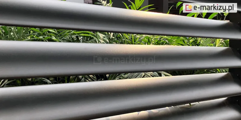 Zdjęcie prezentuje bliski kadr na fragment aluminiowej osłony tarasowej wykonanej z paneli żaluzjowych louvre w kolorze antracytowym. Widoczne są cztery poziome lamele o charakterystycznym, opływowym profilu eliptycznym, zamontowane w równych odstępach. Powierzchnia aluminium posiada matową teksturę, która delikatnie rozprasza naturalne światło słoneczne, podkreślając wypukły kształt elementów. Przez szczeliny pomiędzy lamelami prześwituje gęsta, zielona roślinność ogrodowa, w tym liście palm lub podobnych roślin ozdobnych, co tworzy naturalne tło dla technicznej konstrukcji. Taka budowa panelu zapewnia skuteczne zacienienie i prywatność, nie odcinając całkowicie dopływu światła oraz widoku na otaczającą zieleń.