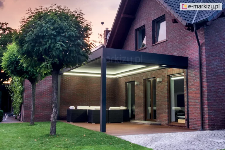 Nowoczesna pergola tarasowa przyścienna zamontowana przy budynku z ciemnoczerwonej cegły klinkierowej. Konstrukcja w kolorze antracytowym posiada zintegrowane oświetlenie LED w formie liniowych pasów zamontowanych wzdłuż wewnętrznych krawędzi ramy, które oświetlają przestrzeń wypoczynkową po zmroku. Pod pergolą, na tarasie wyłożonym deską kompozytową, znajduje się zestaw mebli ogrodowych z technorattanu z jasnymi poduszkami. Obok konstrukcji rosną dwa dekoracyjne drzewa o kulistych koronach, a w tle widoczny jest zadbany trawnik oraz fragment elewacji domu z dużymi przeszkleniami i ciemną dachówką. Zdjęcie prezentuje estetykę i funkcjonalność pergoli bioklimatycznej jako elementu architektury ogrodowej z systemem nocnego podświetlenia.