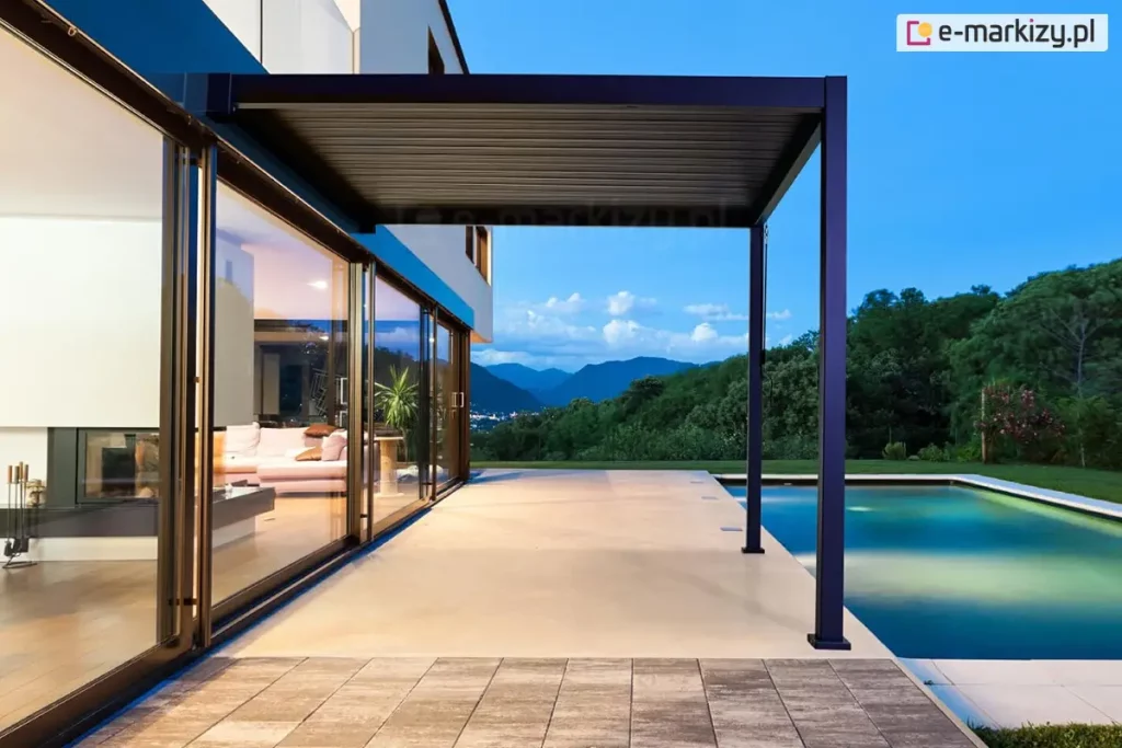 Luksusowa, nowoczesna pergola bioklimatyczna Gloria w kolorze antracytowym, zainstalowana na tarasie przy ekskluzywnej rezydencji. Konstrukcja zadaszenia posiada system ruchomych lameli aluminiowych, które w tej konfiguracji są całkowicie domknięte, tworząc szczelny, płaski dach chroniący przed słońcem lub deszczem. Pergola jest zakotwiczona jedną stroną w elewacji piętrowego budynku, a drugą wspiera się na dwóch smukłych słupach nośnych o kwadratowym przekroju. Taras wykonany jest z gładkich, wielkoformatowych płyt w jasnym kolorze, które kontrastują z pasem ciemniejszego bruku na pierwszym planie. Bezpośrednio przy zadaszonej strefie znajduje się basen typu infinity, w którego tafli wody odbijają się barwy zmierzchu. Budynek posiada panoramiczne, przesuwne przeszklenia, przez które widać nastrojowo oświetlone wnętrze salonu z białą kanapą. Całość osadzona jest w malowniczym krajobrazie z widokiem na zielone wzgórza i góry pod błękitnym niebem w porze błękitnej godziny.