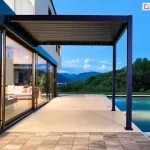 Luksusowa, nowoczesna pergola bioklimatyczna Gloria w kolorze antracytowym, zainstalowana na tarasie przy ekskluzywnej rezydencji. Konstrukcja zadaszenia posiada system ruchomych lameli aluminiowych, które w tej konfiguracji są całkowicie domknięte, tworząc szczelny, płaski dach chroniący przed słońcem lub deszczem. Pergola jest zakotwiczona jedną stroną w elewacji piętrowego budynku, a drugą wspiera się na dwóch smukłych słupach nośnych o kwadratowym przekroju. Taras wykonany jest z gładkich, wielkoformatowych płyt w jasnym kolorze, które kontrastują z pasem ciemniejszego bruku na pierwszym planie. Bezpośrednio przy zadaszonej strefie znajduje się basen typu infinity, w którego tafli wody odbijają się barwy zmierzchu. Budynek posiada panoramiczne, przesuwne przeszklenia, przez które widać nastrojowo oświetlone wnętrze salonu z białą kanapą. Całość osadzona jest w malowniczym krajobrazie z widokiem na zielone wzgórza i góry pod błękitnym niebem w porze błękitnej godziny.