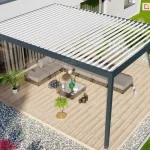 Wizualizacja architektoniczna przedstawiająca nowoczesną pergolę tarasową z dachem lamelowym Bioline, zamontowaną przy ścianie budynku mieszkalnego. Konstrukcja nośna wykonana jest z profili aluminiowych o ciemnoszarym, antracytowym wykończeniu, które harmonijnie współgra z elewacją domu oraz wyposażeniem tarasu. Dach zadaszenia składa się z ciągu równolegle ułożonych, białych ruchomych lameli, które umożliwiają regulację stopnia nasłonecznienia oraz dopływu światła dziennego do przestrzeni wypoczynkowej. Pod pergolą zaaranżowano strefę relaksu z kompletem wypoczynkowym w nowoczesnym stylu, składającym się z modułowej sofy narożnej z szarymi poduszkami, szklanego stolika kawowego oraz dekoracyjnych roślin doniczkowych ustawionych na drewnianej nawierzchni tarasowej. Otoczenie budynku stanowi wypielęgnowany trawnik oraz rabaty żwirowe z ozdobnymi krzewami, co podkreśla estetykę całej posesji. Perspektywa z lotu ptaka ukazuje funkcjonalne wykorzystanie przestrzeni pod zadaszeniem, wskazując na wysoki standard wykończenia oraz spójność designu między architekturą pergoli a zagospodarowaniem ogrodu.