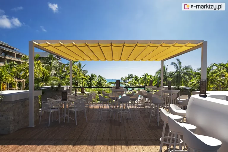 Szeroki kadr prezentujący pergolę rzymską zainstalowaną na tarasie widokowym luksusowego kurortu. Konstrukcja wsparta jest na białych, kwadratowych słupach aluminiowych, a zadaszenie stanowi intensywnie żółta tkanina techniczna, ułożona w symetryczne, głębokie fałdy. Pod pergolą znajduje się strefa kawiarniana wyposażona w nowoczesne, białe meble: wysokie hokery oraz stoliki barowe z kartami menu. Podłoże tarasu wykonane jest z brązowych desek kompozytowych o wyraźnym usłojeniu. W tle rozpościera się egzotyczny krajobraz z licznymi palmami, basenem oraz widokiem na błękitny ocean pod bezchmurnym niebem. Po lewej stronie widoczny jest fragment wielokondygnacyjnego budynku hotelowego o nowoczesnej architekturze. Całość kompozycji podkreśla funkcję ochronną pergoli przed silnym nasłonecznieniem w klimacie tropikalnym.