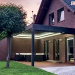 Aluminiowa pergola sb350 selt z podświetleniem w obwodzie dachu na tarasie przy ceglanym domu wieczorową porą. Jasne pióra lameli kontrastują z grafitową konstrukcją