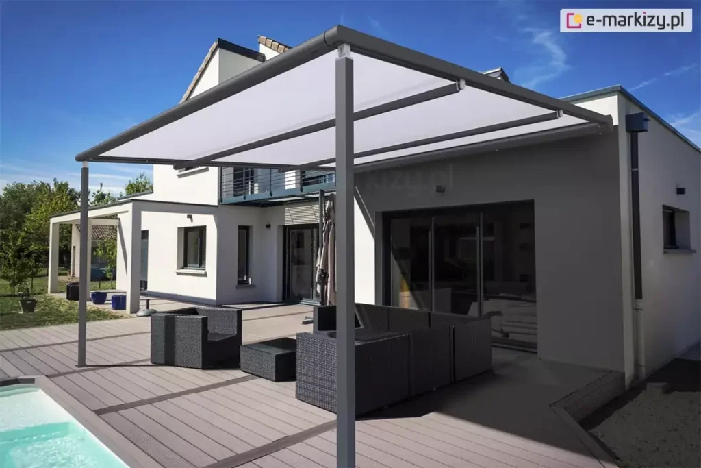 Współczesna, wolnostojąca pergola tarasowa V-Ziiip Aluprof o konstrukcji aluminiowej w kolorze ciemnoszarym (antracytowym), zamontowana na rozległym tarasie z desek kompozytowych. Zadaszenie pergoli stanowi jasna, biała tkanina markizowa, która jest napięta na prowadnicach, tworząc skuteczną osłonę przeciwsłoneczną. Konstrukcja wspiera się na pionowych, kwadratowych słupach. W tle widoczny jest nowoczesny, dwukondygnacyjny dom jednorodzinny o białej elewacji z szarymi wstawkami oraz dużymi przeszkleniami. Na tarasie pod pergolą znajduje się komplet mebli ogrodowych wykonanych z ciemnego technorattanu, składający się z modułowej sofy narożnej, fotela oraz niskiego stolika kawowego. Po lewej stronie zdjęcia widoczny jest fragment krawędzi basenu z błękitną wodą. Otoczenie obejmuje zielony trawnik, drzewa w oddali oraz bezchmurne, niebieskie niebo.