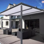 Współczesna, wolnostojąca pergola tarasowa V-Ziiip Aluprof o konstrukcji aluminiowej w kolorze ciemnoszarym (antracytowym), zamontowana na rozległym tarasie z desek kompozytowych. Zadaszenie pergoli stanowi jasna, biała tkanina markizowa, która jest napięta na prowadnicach, tworząc skuteczną osłonę przeciwsłoneczną. Konstrukcja wspiera się na pionowych, kwadratowych słupach. W tle widoczny jest nowoczesny, dwukondygnacyjny dom jednorodzinny o białej elewacji z szarymi wstawkami oraz dużymi przeszkleniami. Na tarasie pod pergolą znajduje się komplet mebli ogrodowych wykonanych z ciemnego technorattanu, składający się z modułowej sofy narożnej, fotela oraz niskiego stolika kawowego. Po lewej stronie zdjęcia widoczny jest fragment krawędzi basenu z błękitną wodą. Otoczenie obejmuje zielony trawnik, drzewa w oddali oraz bezchmurne, niebieskie niebo.