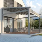 Wolnostojąca pergola tarasowa Elegancy Tarasola z aluminiową konstrukcją i rozwijanym dachem tkaninowym osłania przestronną strefę jadalnianą z metalowymi krzesłami i stolikiem. Nowoczesna forma pergoli podkreśla architekturę budynku, a całość otacza zadbane otoczenie z drzewami i brukowaną nawierzchnią.