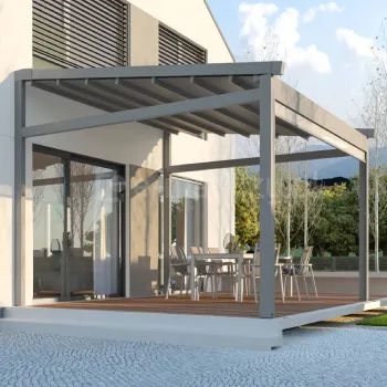 Wolnostojąca pergola tarasowa Elegancy Tarasola z aluminiową konstrukcją i rozwijanym dachem tkaninowym osłania przestronną strefę jadalnianą z metalowymi krzesłami i stolikiem. Nowoczesna forma pergoli podkreśla architekturę budynku, a całość otacza zadbane otoczenie z drzewami i brukowaną nawierzchnią.