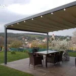 Nowoczesna aluminiowa pergola tarasowa Elegancy Tarasola o konstrukcji w kolorze antracytowym, wyposażona w ruchome zadaszenie tkaninowe w odcieniu kremowym, które jest ułożone w charakterystyczne dla systemu rzymskiego fale. Konstrukcja wsparta na solidnym profilu pionowym jest jednostronnie przytwierdzona do ściany budynku wykończonej dekoracyjnym kamieniem elewacyjnym w odcieniach brązu i szarości. Pod zadaszeniem, na tarasie wyłożonym płytkami imitującymi ciemne drewno, ustawiono zestaw mebli obiadowych z brązowego technorattanu, składający się z prostokątnego stołu z przeszklonym blatem oraz czterech krzeseł. W tle rozpościera się widok na rozległy ogród z zielonym trawnikiem, kwitnącymi na biało drzewami owocowymi oraz panoramiczny krajobraz pagórkowatej okolicy z widoczną w oddali wieżą kościelną pod lekko zachmurzonym niebem.