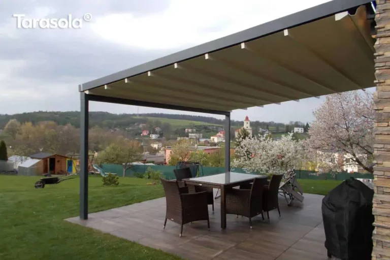 Nowoczesna aluminiowa pergola tarasowa Elegancy Tarasola o konstrukcji w kolorze antracytowym, wyposażona w ruchome zadaszenie tkaninowe w odcieniu kremowym, które jest ułożone w charakterystyczne dla systemu rzymskiego fale. Konstrukcja wsparta na solidnym profilu pionowym jest jednostronnie przytwierdzona do ściany budynku wykończonej dekoracyjnym kamieniem elewacyjnym w odcieniach brązu i szarości. Pod zadaszeniem, na tarasie wyłożonym płytkami imitującymi ciemne drewno, ustawiono zestaw mebli obiadowych z brązowego technorattanu, składający się z prostokątnego stołu z przeszklonym blatem oraz czterech krzeseł. W tle rozpościera się widok na rozległy ogród z zielonym trawnikiem, kwitnącymi na biało drzewami owocowymi oraz panoramiczny krajobraz pagórkowatej okolicy z widoczną w oddali wieżą kościelną pod lekko zachmurzonym niebem.