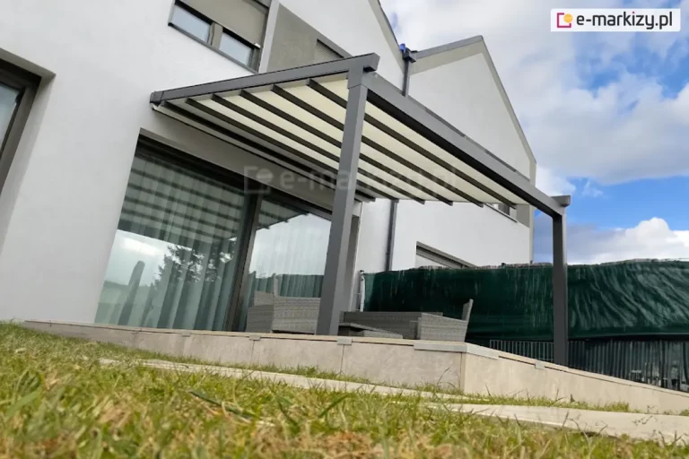 Wizualizacja nowoczesnej pergoli tarasowej o grafitowej, aluminiowej konstrukcji przyściennej, uchwycona w ujęciu od dołu, z perspektywy trawnika. Zadaszenie składa się z kremowego materiału technicznego rozpiętego na równoległych profilach poprzecznych, które są w pełni wysunięte, tworząc lekką, schodkową strukturę. Konstrukcja wspiera się na dwóch solidnych, kwadratowych słupach bocznych o antracytowym wykończeniu. Pod pergolą, na podwyższonym tarasie wyłożonym jasnobeżowymi płytami wielkoformatowymi, widoczny jest zestaw mebli ogrodowych wykonanych z szarego technorattanu. Tło stanowi elewacja nowoczesnego budynku o białym tynku z dużym przeszkleniem drzwi tarasowych, w których odbija się otoczenie. Po prawej stronie taras jest odgrodzony od sąsiedniej posesji za pomocą ciemnozielonej siatki cieniującej zamontowanej na ogrodzeniu. Niebo nad budynkiem jest jasne, z dynamicznie ułożonymi białymi chmurami, co podkreśla nowoczesną architekturę i estetykę instalacji.