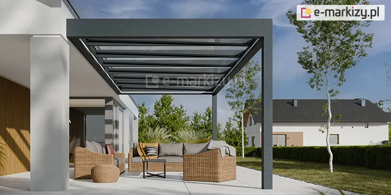 Wizualizacja przedstawia aluminiowe zadaszenie tarasowe model Spekta w wersji przyściennej, charakteryzujące się minimalistycznym, kubistycznym designem. Konstrukcja wykonana jest z grubych profili o prostokątnym przekroju w kolorze antracytowym, z wyraźnie zaznaczoną wysoką ramą frontową, która maskuje spadek dachu. Zadaszenie wspiera się na dwóch solidnych słupach bocznych i jest zamontowane bezpośrednio do białej elewacji nowoczesnego budynku. Dach wypełniają podłużne panele szklane, pod którymi widoczne są zintegrowane listwy oświetleniowe LED, biegnące wzdłuż całego obwodu zadaszenia. Pod wiatą zaaranżowano przestronną strefę relaksu z meblami z technorattanu w naturalnym, słomkowym kolorze, wyposażonymi w szare poduszki siedziskowe. Zestaw składa się z narożnej sofy, fotela oraz plecionego pufa, ustawionych wokół małego stolika kawowego z czarnym blatem. Taras wyłożony jest dużymi, jasnoszarymi płytami. W tle widoczny jest zielony ogród z drzewami liściastymi oraz fragment sąsiedniego domu o białej elewacji i ciemnym dachu, co podkreśla podmiejski charakter inwestycji.