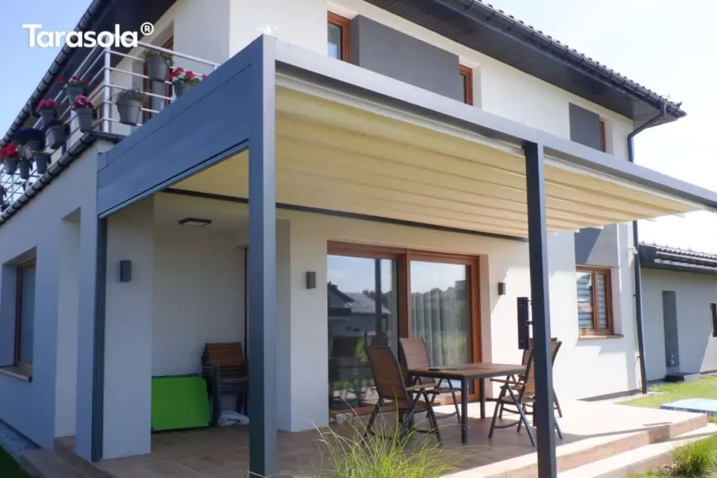 Zdjęcie prezentuje przyścienną pergolę tarasową Tarasola Flat o konstrukcji aluminiowej w kolorze ciemnego antracytu, zamontowaną na nowoczesnym budynku jednorodzinnym. Zadaszenie stanowi jasny, kremowy materiał ułożony w charakterystyczne, poziome fałdy, co wskazuje na system pergoli segmentowej (tkaninowej). Konstrukcja wspiera się na solidnych pionowych słupach o kwadratowym przekroju, a rynna zintegrowana z profilem bocznym po lewej stronie służy do odprowadzania wody opadowej. Pod pergolą znajduje się wykafelkowany taras, na którym ustawiono drewniany stół z kompletem krzeseł. Elewacja budynku jest biała z szarymi wstawkami wokół okien oraz drewnianą stolarką okienną. Na piętrze widoczny jest balkon z metalową barierką, ozdobiony donicami z czerwonymi kwiatami. Całość aranżacji dopełniają nowoczesne kinkiety zewnętrzne zamontowane na ścianie budynku oraz niska roślinność ogrodowa widoczna w dolnej części kadru.