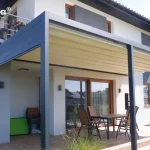 Zdjęcie prezentuje przyścienną pergolę tarasową Tarasola Flat o konstrukcji aluminiowej w kolorze ciemnego antracytu, zamontowaną na nowoczesnym budynku jednorodzinnym. Zadaszenie stanowi jasny, kremowy materiał ułożony w charakterystyczne, poziome fałdy, co wskazuje na system pergoli segmentowej (tkaninowej). Konstrukcja wspiera się na solidnych pionowych słupach o kwadratowym przekroju, a rynna zintegrowana z profilem bocznym po lewej stronie służy do odprowadzania wody opadowej. Pod pergolą znajduje się wykafelkowany taras, na którym ustawiono drewniany stół z kompletem krzeseł. Elewacja budynku jest biała z szarymi wstawkami wokół okien oraz drewnianą stolarką okienną. Na piętrze widoczny jest balkon z metalową barierką, ozdobiony donicami z czerwonymi kwiatami. Całość aranżacji dopełniają nowoczesne kinkiety zewnętrzne zamontowane na ścianie budynku oraz niska roślinność ogrodowa widoczna w dolnej części kadru.