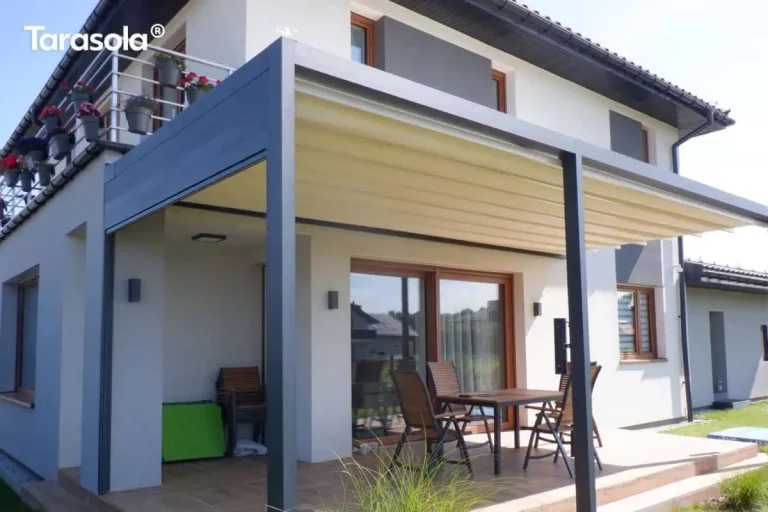 Zdjęcie prezentuje przyścienną pergolę tarasową Tarasola Flat o konstrukcji aluminiowej w kolorze ciemnego antracytu, zamontowaną na nowoczesnym budynku jednorodzinnym. Zadaszenie stanowi jasny, kremowy materiał ułożony w charakterystyczne, poziome fałdy, co wskazuje na system pergoli segmentowej (tkaninowej). Konstrukcja wspiera się na solidnych pionowych słupach o kwadratowym przekroju, a rynna zintegrowana z profilem bocznym po lewej stronie służy do odprowadzania wody opadowej. Pod pergolą znajduje się wykafelkowany taras, na którym ustawiono drewniany stół z kompletem krzeseł. Elewacja budynku jest biała z szarymi wstawkami wokół okien oraz drewnianą stolarką okienną. Na piętrze widoczny jest balkon z metalową barierką, ozdobiony donicami z czerwonymi kwiatami. Całość aranżacji dopełniają nowoczesne kinkiety zewnętrzne zamontowane na ścianie budynku oraz niska roślinność ogrodowa widoczna w dolnej części kadru.