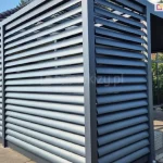 Zdjęcie przedstawia nowoczesną pergolę tarasową o konstrukcji aluminiowej w kolorze antracytowym, wyposażoną w stałe panele żaluzjowe typu louvre. Jedna ściana boczna pergoli została w pełni wypełniona poziomymi lamelami o eliptycznym przekroju, które są zamontowane pod stałym kątem, tworząc szczelną, a jednocześnie ażurową przesłonę. Osłonę panelem żaluzjowym Louvre wykorzystano także na części jednej z bocznych ścian pergoli. Konstrukcja opiera się na solidnych, kwadratowych profilach pionowych, a całość jest posadowiona na betonowym tarasie wyłożonym dużymi płytami. W tle widoczny jest fragment budynku mieszkalnego z jasną elewacją i ciemnym dachem oraz przylegający ogród z zieloną roślinnością. Zastosowane panele żaluzjowe pełnią funkcję estetycznej zabudowy bocznej, zapewniając ochronę przed słońcem, wiatrem oraz gwarantując prywatność osobom przebywającym pod zadaszeniem.
