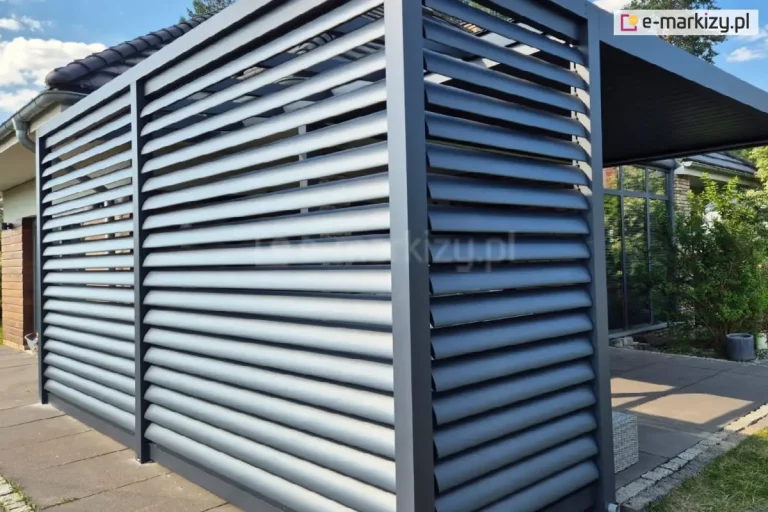 Zdjęcie przedstawia nowoczesną pergolę tarasową o konstrukcji aluminiowej w kolorze antracytowym, wyposażoną w stałe panele żaluzjowe typu louvre. Jedna ściana boczna pergoli została w pełni wypełniona poziomymi lamelami o eliptycznym przekroju, które są zamontowane pod stałym kątem, tworząc szczelną, a jednocześnie ażurową przesłonę. Osłonę panelem żaluzjowym Louvre wykorzystano także na części jednej z bocznych ścian pergoli. Konstrukcja opiera się na solidnych, kwadratowych profilach pionowych, a całość jest posadowiona na betonowym tarasie wyłożonym dużymi płytami. W tle widoczny jest fragment budynku mieszkalnego z jasną elewacją i ciemnym dachem oraz przylegający ogród z zieloną roślinnością. Zastosowane panele żaluzjowe pełnią funkcję estetycznej zabudowy bocznej, zapewniając ochronę przed słońcem, wiatrem oraz gwarantując prywatność osobom przebywającym pod zadaszeniem.