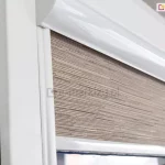 Zbliżenie na górną część białej ramy okiennej z zamontowaną kasetową roletą wewnętrzną. Centralnym punktem zdjęcia jest materiał rolety o wyraźnej, poziomej strukturze przypominającej plecionkę lub naturalne włókna w odcieniach beżu, brązu i szarości. Tkanina jest częściowo rozwinięta i chowa się wewnątrz białej, lakierowanej kasety o zaokrąglonym profilu, która jest zamontowana u góry ramy okna. Po lewej stronie widoczna jest pionowa, biała prowadnica boczna, która przylega bezpośrednio do profilu okiennego i utrzymuje materiał rolety blisko szyby. Dolna krawędź widocznego fragmentu rolety zakończona jest estetyczną, białą listwą obciążającą. Cała konstrukcja rolety oraz rama okna są wykonane w kolorze białym o wysokim połysku, co kontrastuje z matową i fakturowaną strukturą tkaniny przesłonowej. Światło padające na zestaw podkreśla detale splotu materiału oraz gładką powierzchnię elementów aluminiowych.