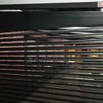 Zdjęcie prezentuje aluminiową ściankę żaluzjową Louvre zamontowaną jako boczny panel wypełniający w konstrukcji pergoli tarasowej. Przesłona składa się z licznych, równolegle ułożonych poziomych lameli o obłym, eliptycznym przekroju w kolorze głębokiego antracytu. Górna część panelu została wykończona szerokim, pełnym profilem maskującym o trapezowym kształcie, który idealnie dopasowuje się do kąta nachylenia dachu pergoli. Całość konstrukcji jest osadzona w pionowych prowadnicach, a po lewej stronie widoczny jest fragment elewacji budynku wykonanej z czerwonej cegły. Przez ażurowe odstępy między lamelami prześwituje wnętrze tarasu, tworząc estetyczną barierę wizualną, która zapewnia prywatność przy zachowaniu cyrkulacji powietrza.