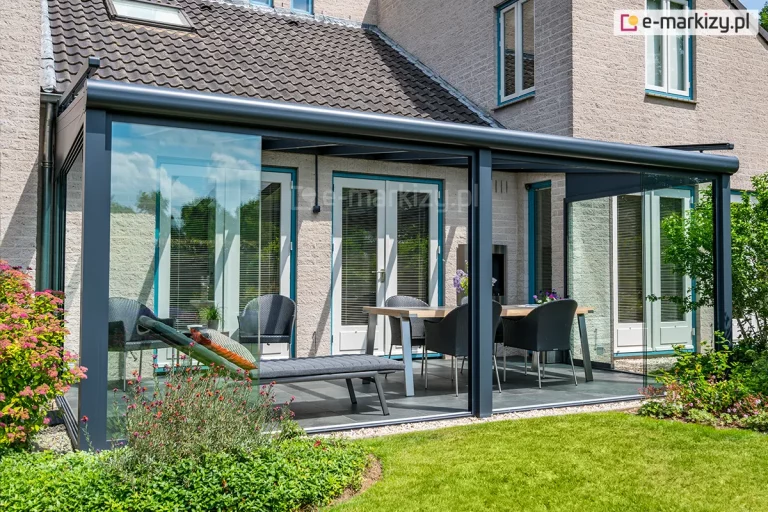 Nowoczesna zabudowa tarasu w formie ogrodu letniego, oparta na aluminiowej konstrukcji w kolorze antracytowym. Zadaszenie jest zamontowane do jasnej, tynkowanej elewacji budynku i posiada dach wykonany z przezroczystych tafli szkła. Frontowa oraz boczna ściana konstrukcji zostały wyposażone w system bezramowych drzwi przesuwnych ze szkła hartowanego, które poruszają się po wielotorowych szynach dolnych. Jeden z paneli frontowych jest częściowo odsunięty, co obrazuje funkcjonalność systemu. Pod zadaszeniem znajduje się strefa wypoczynkowa z szarym szezlongiem z dekoracyjną poduszką oraz strefa jadalniana z drewnianym stołem i czarnymi fotelami z technorattanu. W tle widoczne są białe drzwi tarasowe z pionowymi żaluzjami wewnętrznymi. Na zewnątrz zadaszenia, wzdłuż jego krawędzi, wysypano pas jasnego żwiru, który graniczy z zadbanym trawnikiem. Po lewej stronie kadru widoczne są kwitnące krzewy ogrodowe oraz fragment dachu sąsiedniego budynku krytego ciemną dachówką.