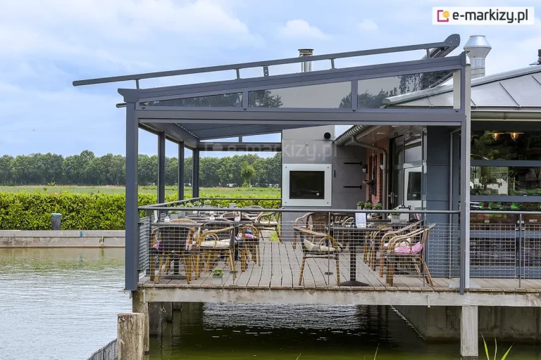 Widok z boku na komercyjne zadaszenie tarasu Terano posadowione na betonowych filarach nad wodą. Konstrukcja w kolorze antracytowym posiada boczny klin szklany o kształcie trapezu, który wypełnia przestrzeń między dachem a balustradą. Na drewnianym pomoście tarasowym ustawiono zestawy kawiarniane. Dach konstrukcji jest jednospadowy, a w tle widać otwarty krajobraz z linią drzew i zbiornikiem wodnym.