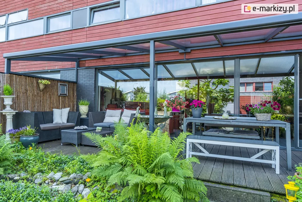 Zdjęcie prezentuje nowoczesne, przyścienne zadaszenie tarasowe nebbiolo deponti o konstrukcji aluminiowej w kolorze antracytowym, zintegrowane z elewacją budynku wykończoną panelami w kolorze ceglastym. Dach konstrukcji wykonany jest z przezroczystych płyt poliwęglanowych, które wpuszczają naturalne światło, chroniąc jednocześnie przestrzeń przed opadami. Pod zadaszeniem zaaranżowano bogatą strefę wypoczynkową, obejmującą narożną sofę z technorattanu z jasnymi poduszkami, duży stół jadalniany z ławami oraz liczne rośliny ozdobne w donicach, w tym intensywnie różowe pelargonie i paprocie. Taras wyłożony jest ciemnymi deskami kompozytowymi, a jego krawędź sąsiaduje z pasem dekoracyjnych kamieni i gęstą roślinnością ogrodową. Przeszklone, przesuwne drzwi tarasowe odbijają otoczenie ogrodu, tworząc wrażenie płynnego przejścia między wnętrzem domu a przestrzenią zewnętrzną.