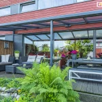 Zdjęcie prezentuje nowoczesne, przyścienne zadaszenie tarasowe nebbiolo deponti o konstrukcji aluminiowej w kolorze antracytowym, zintegrowane z elewacją budynku wykończoną panelami w kolorze ceglastym. Dach konstrukcji wykonany jest z przezroczystych płyt poliwęglanowych, które wpuszczają naturalne światło, chroniąc jednocześnie przestrzeń przed opadami. Pod zadaszeniem zaaranżowano bogatą strefę wypoczynkową, obejmującą narożną sofę z technorattanu z jasnymi poduszkami, duży stół jadalniany z ławami oraz liczne rośliny ozdobne w donicach, w tym intensywnie różowe pelargonie i paprocie. Taras wyłożony jest ciemnymi deskami kompozytowymi, a jego krawędź sąsiaduje z pasem dekoracyjnych kamieni i gęstą roślinnością ogrodową. Przeszklone, przesuwne drzwi tarasowe odbijają otoczenie ogrodu, tworząc wrażenie płynnego przejścia między wnętrzem domu a przestrzenią zewnętrzną.