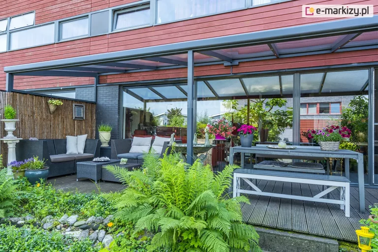 Zdjęcie prezentuje nowoczesne, przyścienne zadaszenie tarasowe nebbiolo deponti o konstrukcji aluminiowej w kolorze antracytowym, zintegrowane z elewacją budynku wykończoną panelami w kolorze ceglastym. Dach konstrukcji wykonany jest z przezroczystych płyt poliwęglanowych, które wpuszczają naturalne światło, chroniąc jednocześnie przestrzeń przed opadami. Pod zadaszeniem zaaranżowano bogatą strefę wypoczynkową, obejmującą narożną sofę z technorattanu z jasnymi poduszkami, duży stół jadalniany z ławami oraz liczne rośliny ozdobne w donicach, w tym intensywnie różowe pelargonie i paprocie. Taras wyłożony jest ciemnymi deskami kompozytowymi, a jego krawędź sąsiaduje z pasem dekoracyjnych kamieni i gęstą roślinnością ogrodową. Przeszklone, przesuwne drzwi tarasowe odbijają otoczenie ogrodu, tworząc wrażenie płynnego przejścia między wnętrzem domu a przestrzenią zewnętrzną.